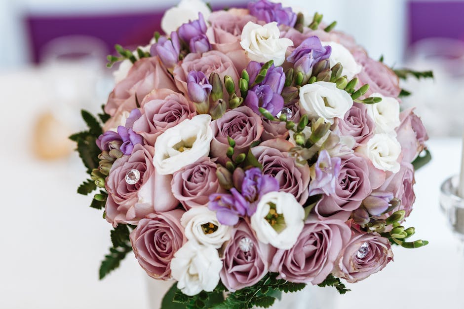 A close-up view of a floral arrangement featuring a bouquet of fresh, pastel pink roses, white lisianthus, and purple freesia, interspersed with green foliage. The bouquet is arranged in a rounded sha