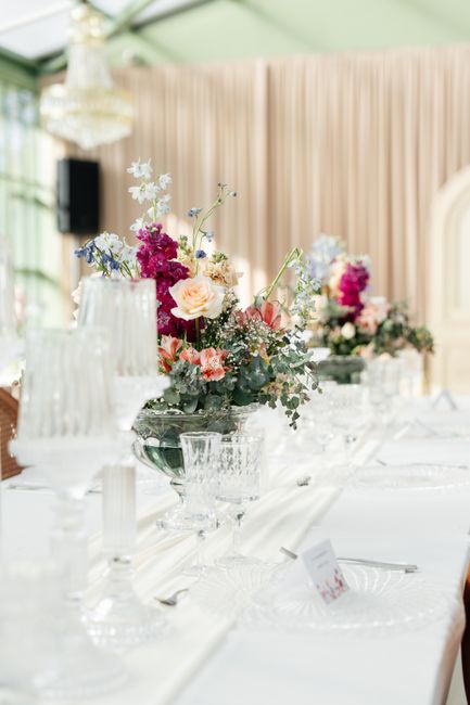 A floral arrangement featuring a mix of pastel and vibrant flowers including pale pink roses, white and blue delphiniums, and deep magenta blossoms, set in a clear glass vase with a textured pattern. 