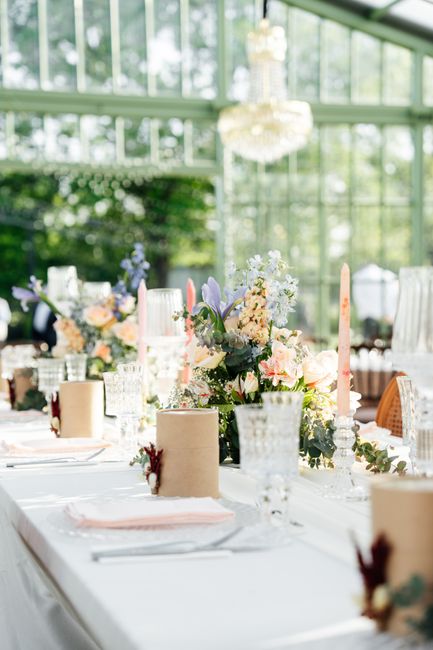 A floral arrangement featuring a variety of fresh flowers, including pale pink roses, purple irises, and delicate white blooms, is displayed atop a white tablecloth during a wedding reception. The bou