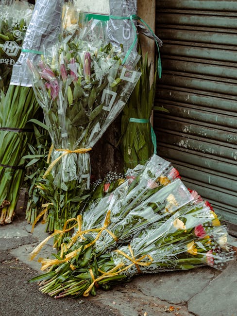 Several bouquets of fresh flowers, including pink lilies, roses, and mixed greenery, wrapped in clear cellophane and tied with yellow and green ribbons, are displayed on the pavement outside a shop wi