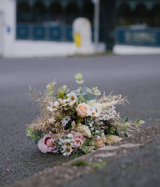 A delicate bouquet of fresh flowers resting on the pavement near a kerb, featuring pale pink roses, white daisies, and small white filler flowers complemented by soft greenery and wispy, dried foliage